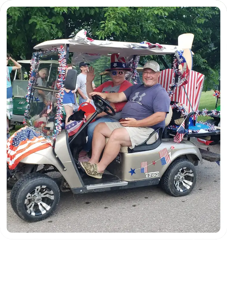 A smiling man and woman riding in an E-Z-GO golf cart decorated with American flags, tinsel, and patriotic banners for an event at Kountry Resort Campground.
