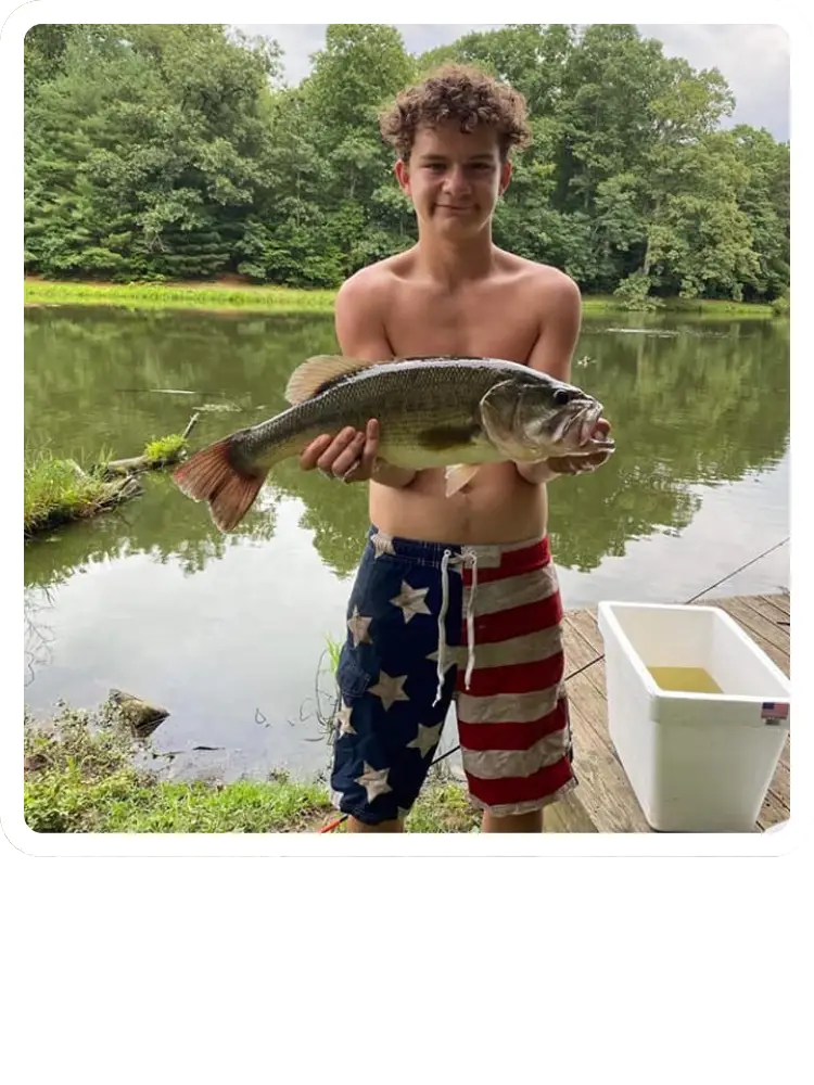 A teenage boy in American flag swim trunks standing on a wooden dock and proudly holding up a large largemouth bass he caught at Kountry Resort Campground.