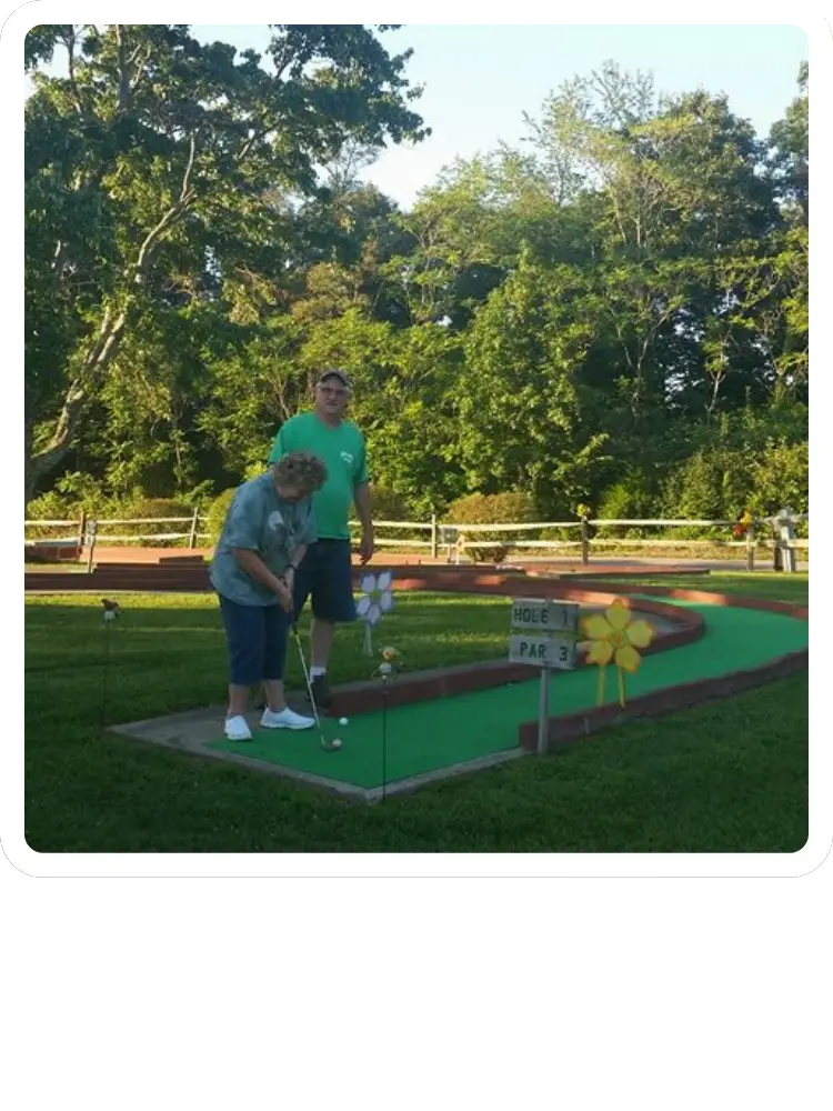 A woman preparing to putt a golf ball on a green mini-golf course while a man watches on a sunny afternoon at Kountry Resort Campground.