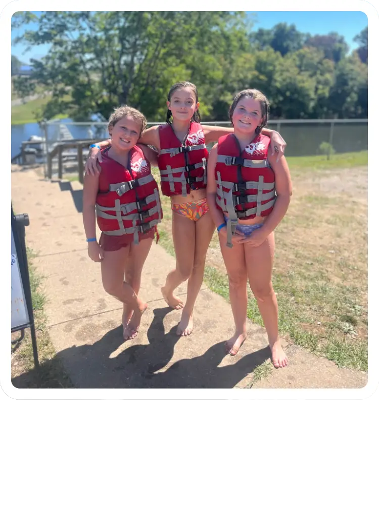 Three young girls smiling and posing together in red life jackets on a sunny walkway near the lake at Kountry Resort Campground.