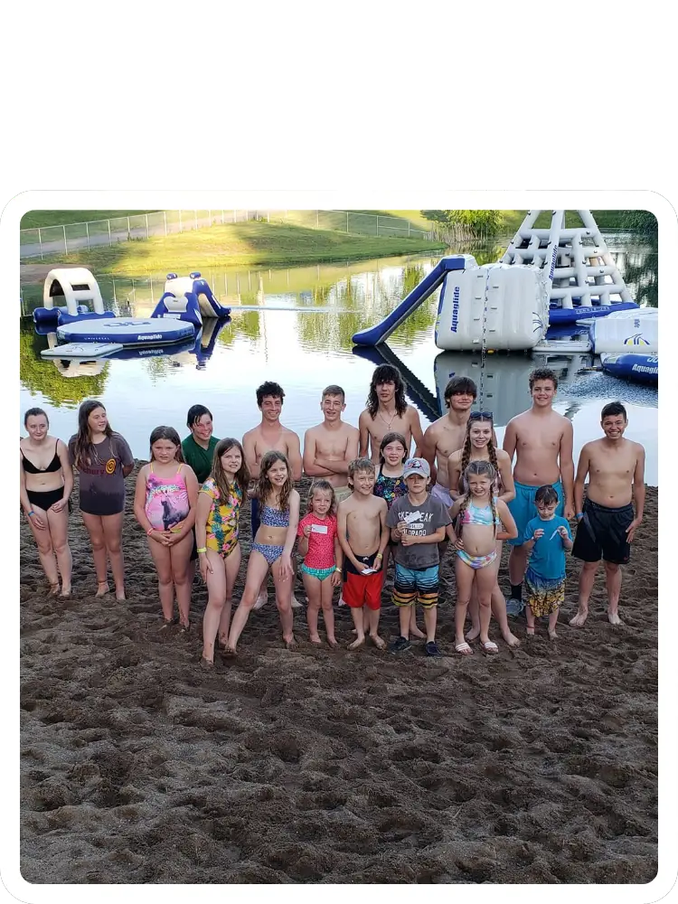 A large group of children and teenagers in swimwear smiling together on the sandy beach in front of the floating lake Aqua Park at Kountry Resort Campground.