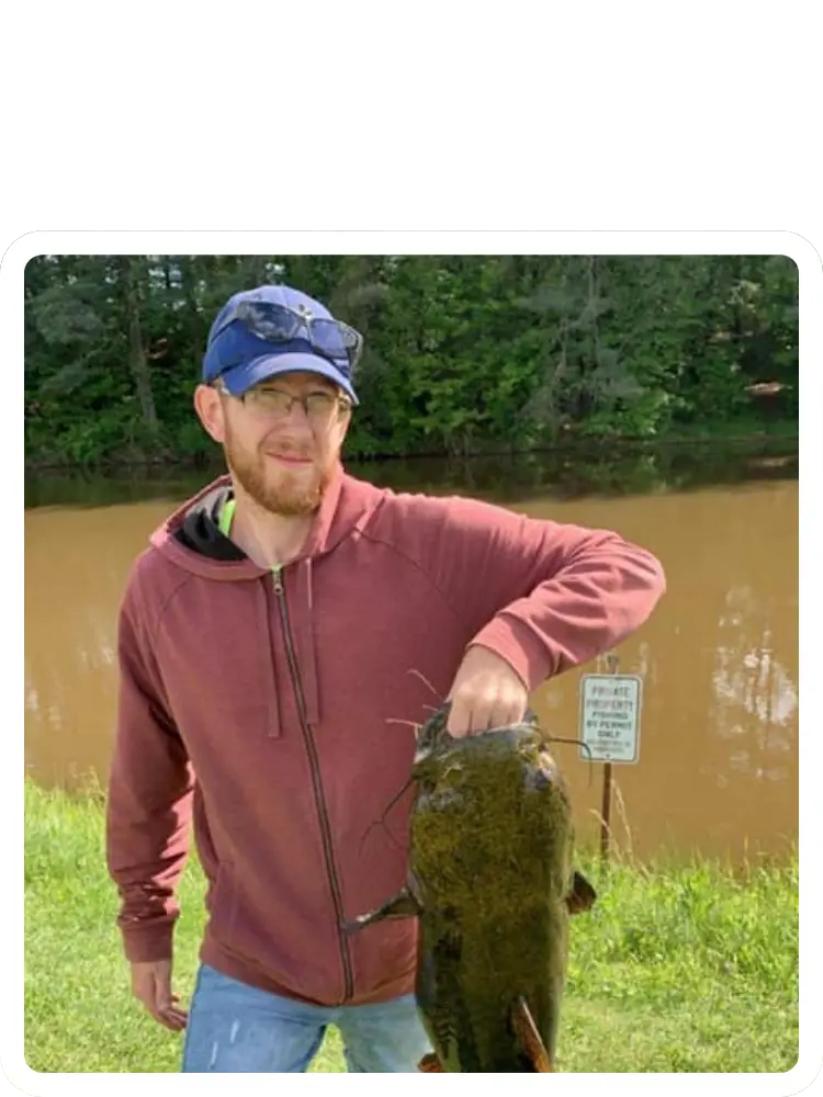A fisherman wearing a blue cap and a maroon hoodie holding up a large catfish by the mouth at the edge of a lake at Kountry Resort Campground.