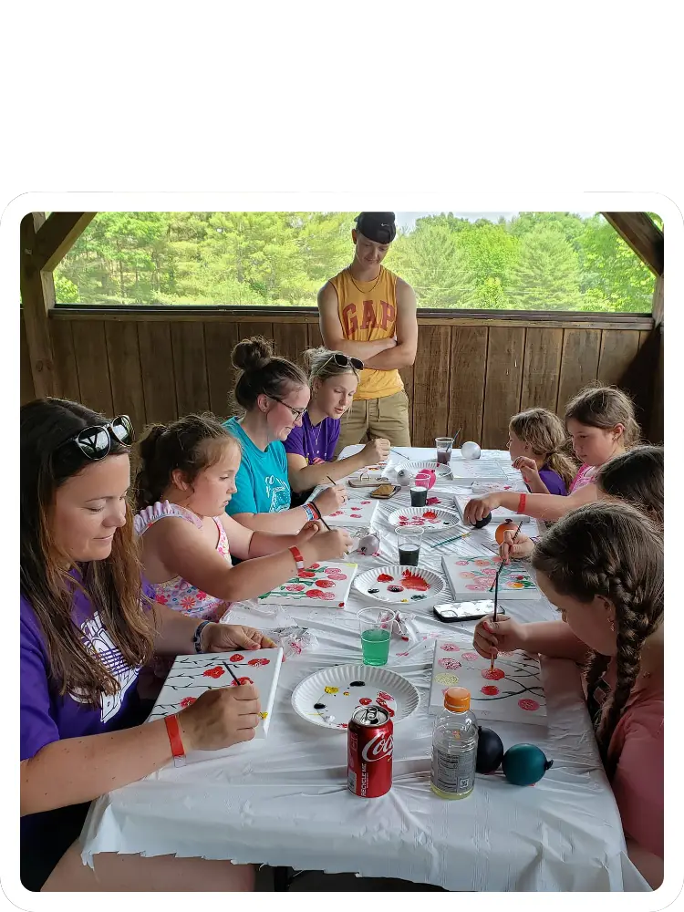 A group of children and adults sitting along a long table inside an open-air pavilion, painting floral designs on small canvases during an activities session at Kountry Resort Campground.