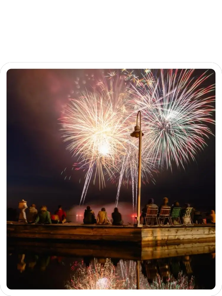 A group of spectators sitting on a wooden lakeside dock watching a vibrant colorful fireworks display overhead at Kountry Resort Campground.