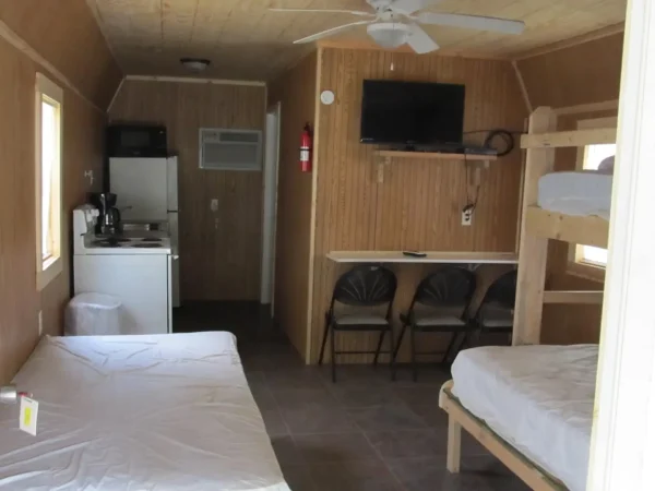 The wood-paneled interior of a rental cabin featuring a small kitchen area, a dining counter with chairs, a wall-mounted TV, a single bed, and wooden bunk beds at Kountry Resort Campground.