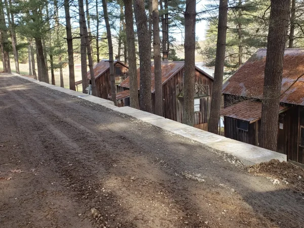 A view looking down a dirt access road alongside a newly constructed retaining wall, overlooking a row of rustic wooden cabins nestled in a pine forest at Kountry Resort Campground.