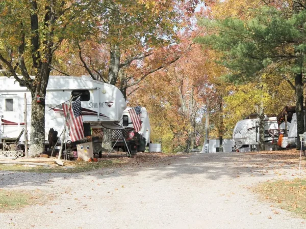 A view looking down a gravel campground road lined with white fifth-wheel trailers decorated with American flags, surrounded by trees displaying colorful fall foliage at Kountry Resort Campground.