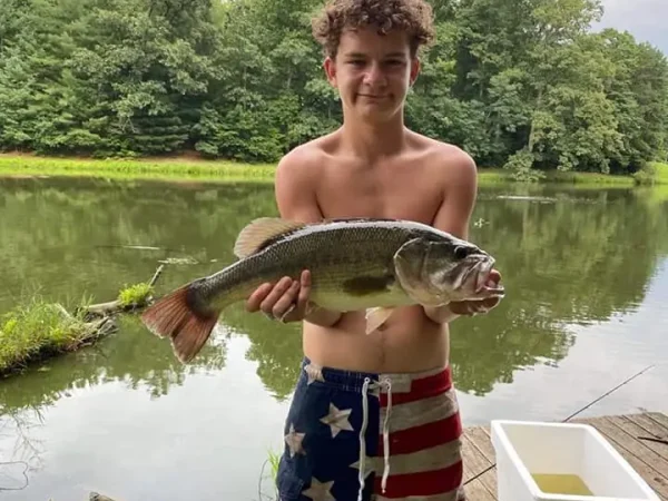 A teenage boy wearing American flag swim shorts standing by a lake shoreline and proudly holding up a large largemouth bass at Kountry Resort Campground.