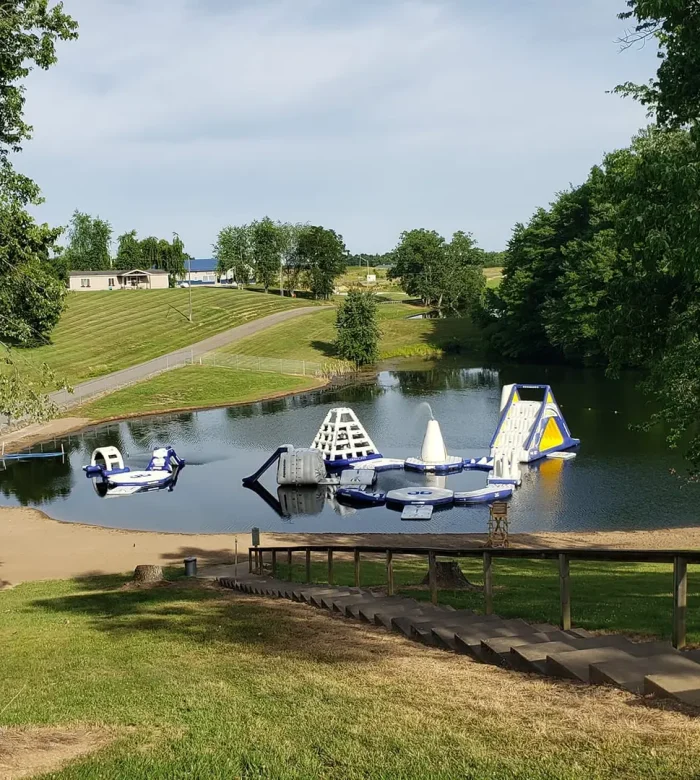 A scenic, distant high-angle view of the lakefront Aqua Park and beach area at Kountry Resort Campground, framed by grassy slopes and trees.