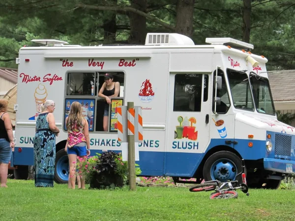 A classic white and blue Mister Softee ice cream truck parked on a grassy lawn with guests gathered at the order window at Kountry Resort Campground.