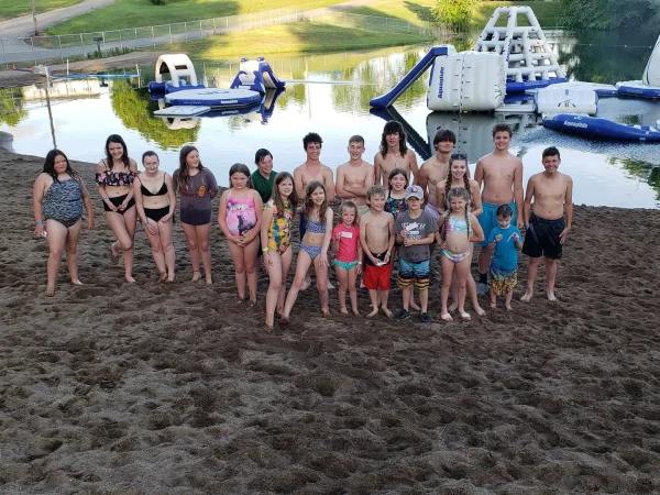A large group of children and teenagers in swimwear standing together on a sandy beach in front of a lake with an inflatable aqua park obstacle course.