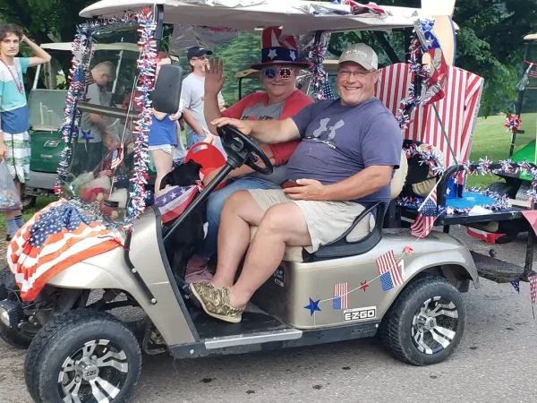A man and a woman smiling and waving from a gold E-Z-GO golf cart decorated with patriotic red, white, and blue tinsel and American flags at Kountry Resort Campground.