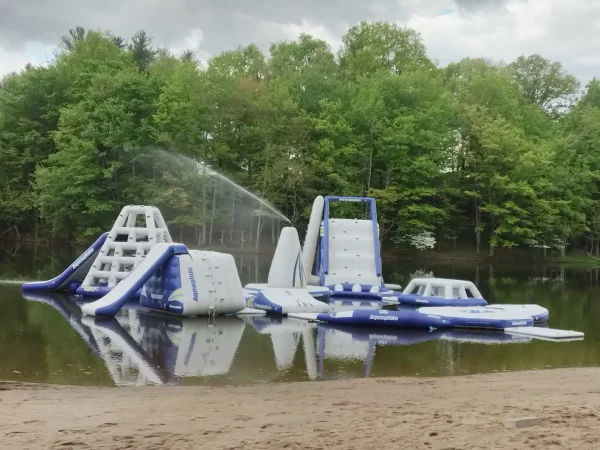 A wide view of a large, interconnected blue and white Aquaglide inflatable water park obstacle course floating on a calm lake surface next to a sandy beach at Kountry Resort Campground.
