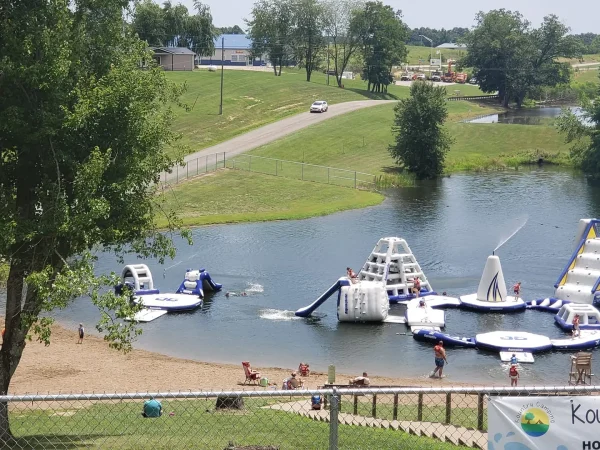 High-angle view of the large lakefront Aqua Park at Kountry Resort Campground, featuring inflatable water slides, trampolines, and climbing structures near a sandy beach.