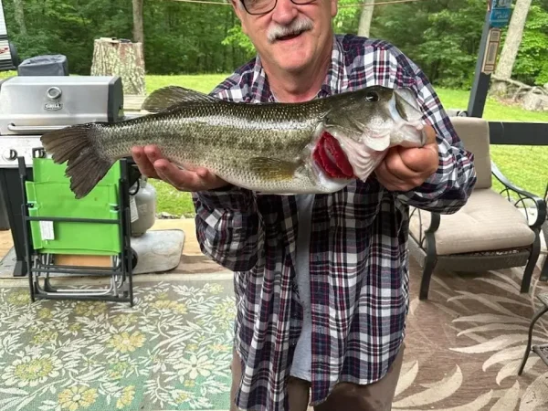 A smiling older man with glasses, a mustache, and a plaid shirt standing on a covered patio while holding up a very large largemouth bass at Kountry Resort Campground.