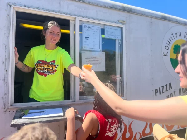 A smiling food truck vendor in a neon yellow shirt handing a cup of food through a service window to guests at Kountry Resort Campground.