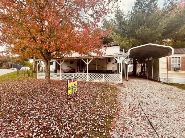 A permanent park model travel trailer with a large covered front porch, white lattice fencing, and an adjacent metal carport setup at Kountry Resort Campground.