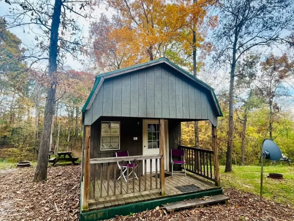 A cozy dark-painted wooden rental cabin with a covered front porch, patio chairs, and a gambrel roof surrounded by fall foliage at Kountry Resort Campground.