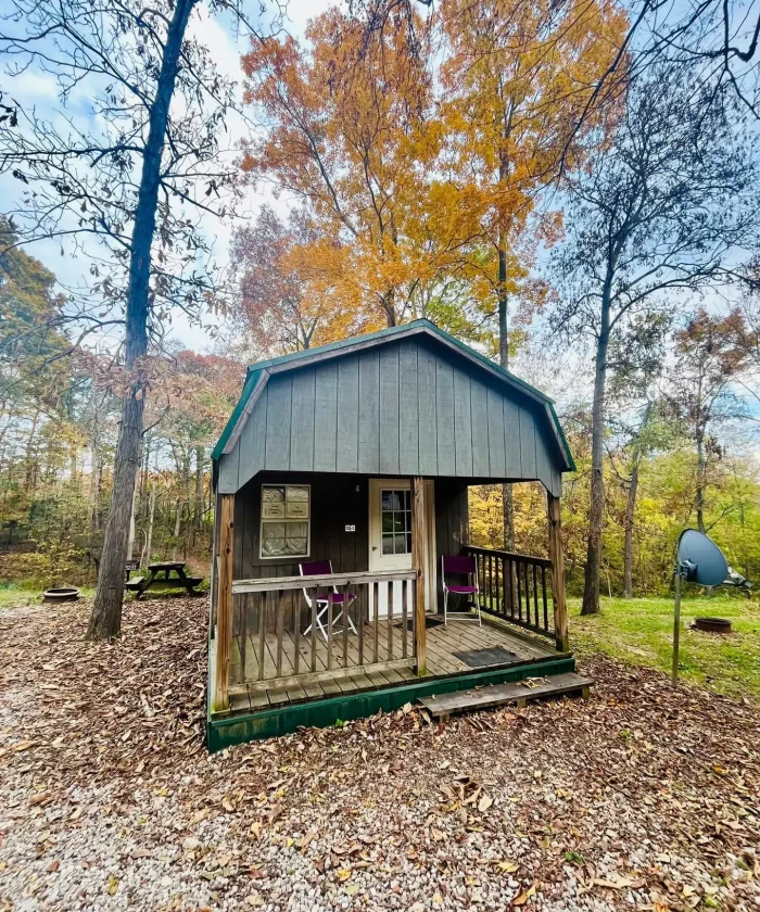 A cozy dark-painted wooden rental cabin with a covered front porch, patio chairs, and a gambrel roof surrounded by fall foliage at Kountry Resort Campground.