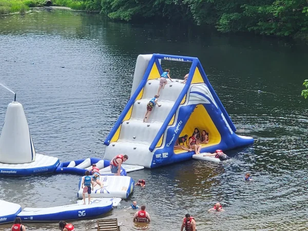 Children and adults wearing life jackets playing on a large blue, white, and yellow inflatable water slide structure in a lake at Kountry Resort Campground.