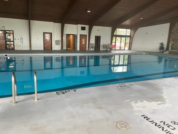 A wide view of the indoor swimming pool deck showing the pool ladders, depth markers, and changing room entryways under a vaulted wood ceiling at Kountry Resort Campground.
