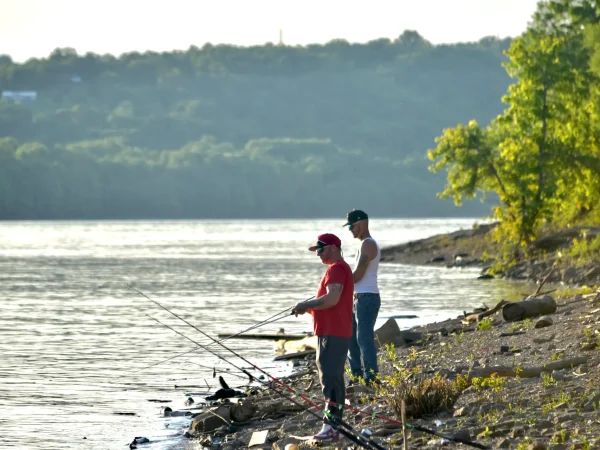 Two men standing on a rocky shoreline holding fishing rods while angling in a wide river under late afternoon light at Kountry Resort Campground.