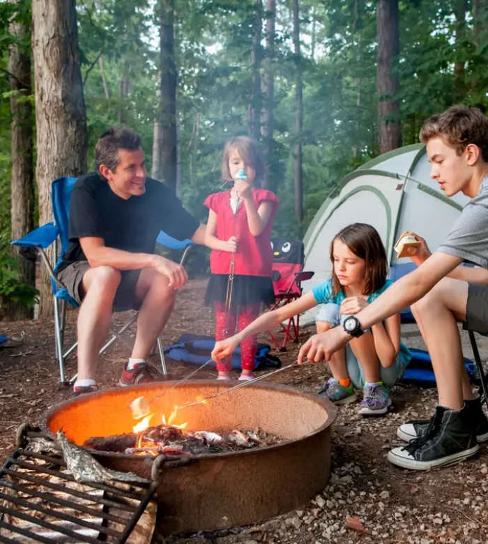 A family sitting in folding chairs around a glowing metal fire ring, roasting marshmallows in front of their tent at Kountry Resort Campground.