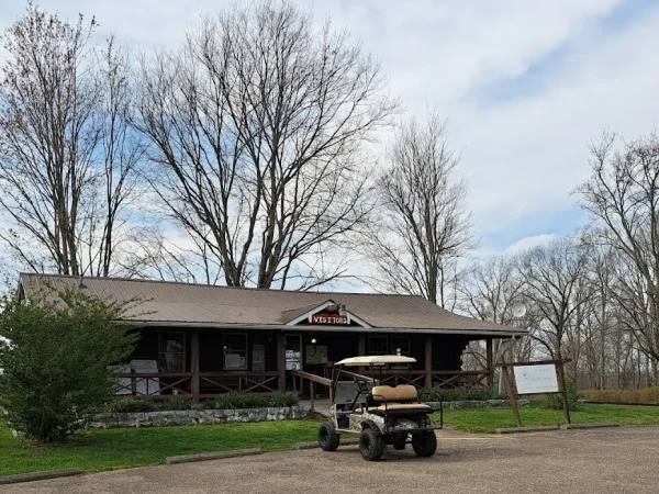 A single-story wooden log cabin check-in office with a front porch, a parked golf cart in a gravel parking lot, and tall trees under a cloudy sky at Kountry Resort Campground.