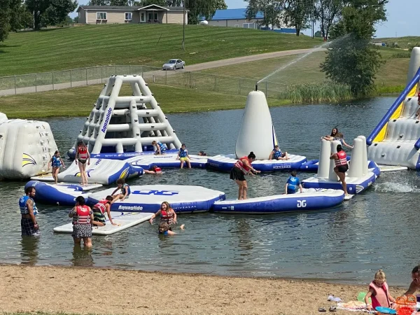 Children and adults wearing life jackets playing on an inflatable water park obstacle course with climbing towers and slides in a lake at Kountry Resort Campground.