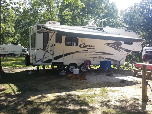 A white Keystone Cougar fifth-wheel travel trailer with its awning extended at a shaded campground site at Kountry Resort Campground.