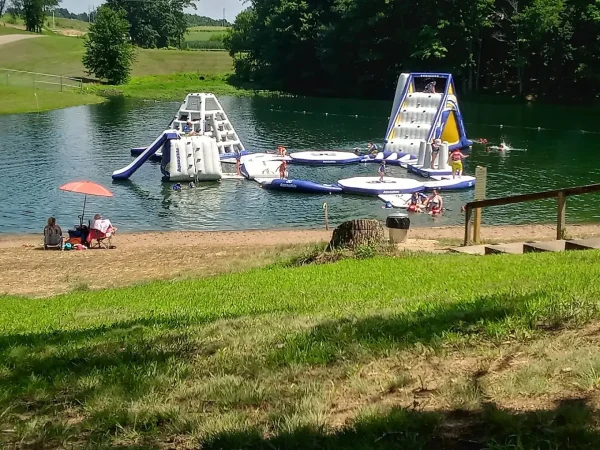 A floating blue and white inflatable water park obstacle course in a lake with people relaxing on a sandy beach under an umbrella at Kountry Resort Campground.