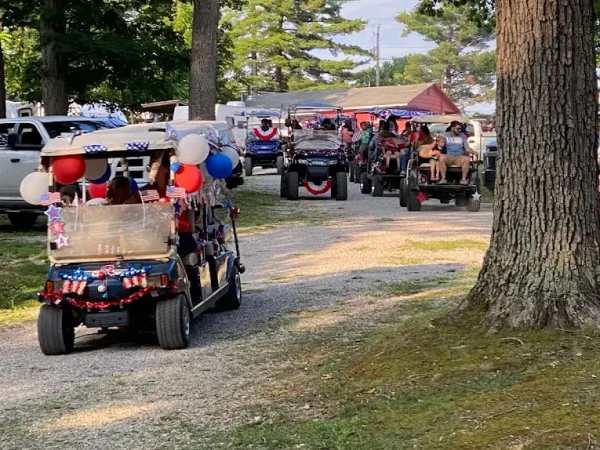A line of golf carts decorated with patriotic red, white, and blue balloons and flags driving along a gravel camp lane during a parade at Kountry Resort Campground.