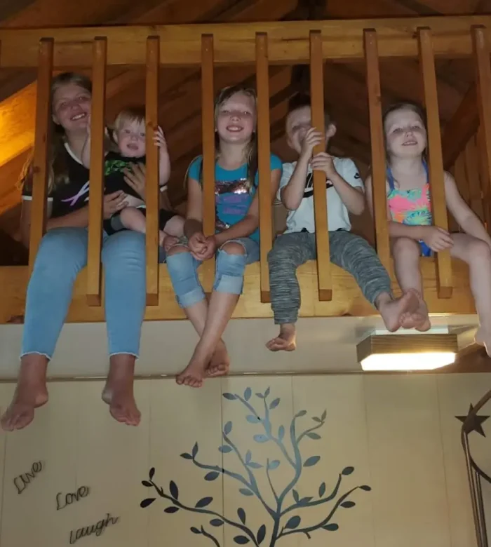 A group of five smiling children sitting on the edge of a wooden cabin loft railing inside a cozy vacation rental at Kountry Resort Campground.