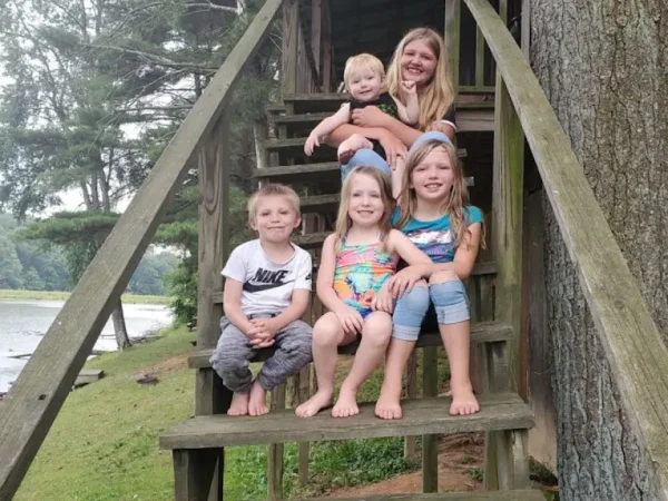 A group of five children smiling and posing together on the wooden exterior steps of a rental cabin near a lake at Kountry Resort Campground.