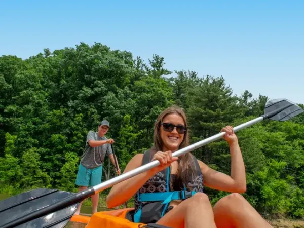 A smiling young woman in sunglasses kayaking in a bright orange boat with a man paddleboarding in the background at Kountry Resort Campground.