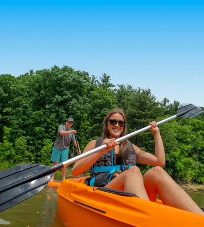 A smiling young woman in sunglasses kayaking in a bright orange boat with a man paddleboarding in the background at Kountry Resort Campground.