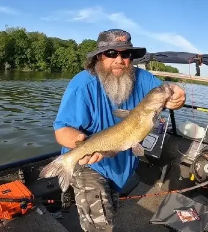 A bearded fisherman wearing a sun hat and sunglasses proudly holding up a large channel catfish on his boat at Kountry Resort Campground.