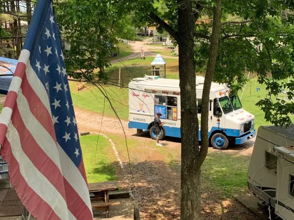 An American flag hanging in the foreground with a classic white and blue ice cream truck parked on a dirt path under green trees at Kountry Resort Campground.