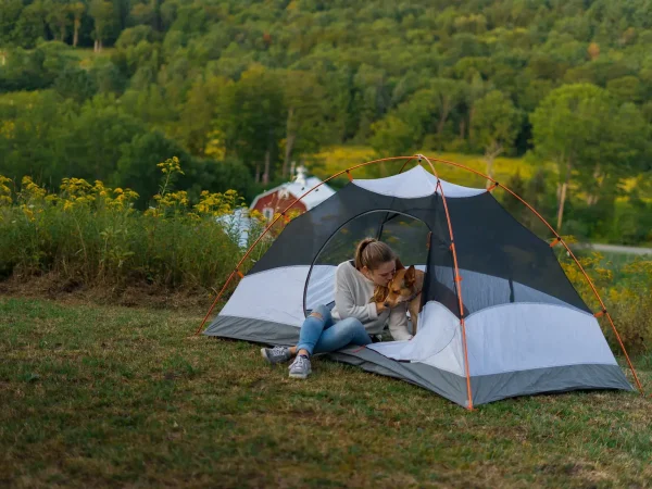 A young woman sitting inside a small dome tent pitching on a grassy ridge while petting her dog, overlooking rolling green hills and a distant barn at Kountry Resort Campground.